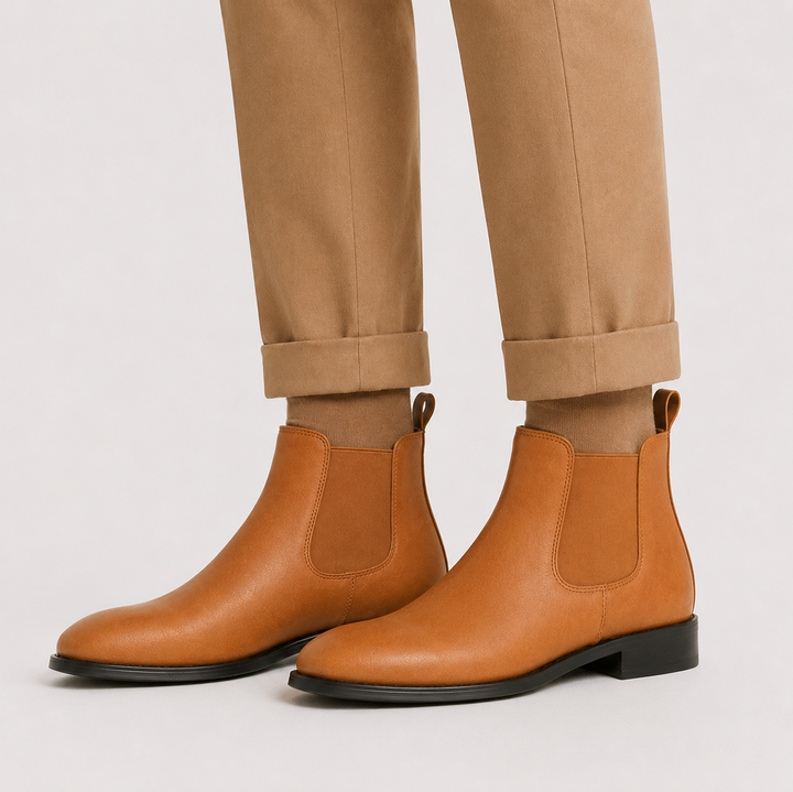 Tan leather Chelsea ankle boots worn with cuffed tan trousers against a light gray studio background.