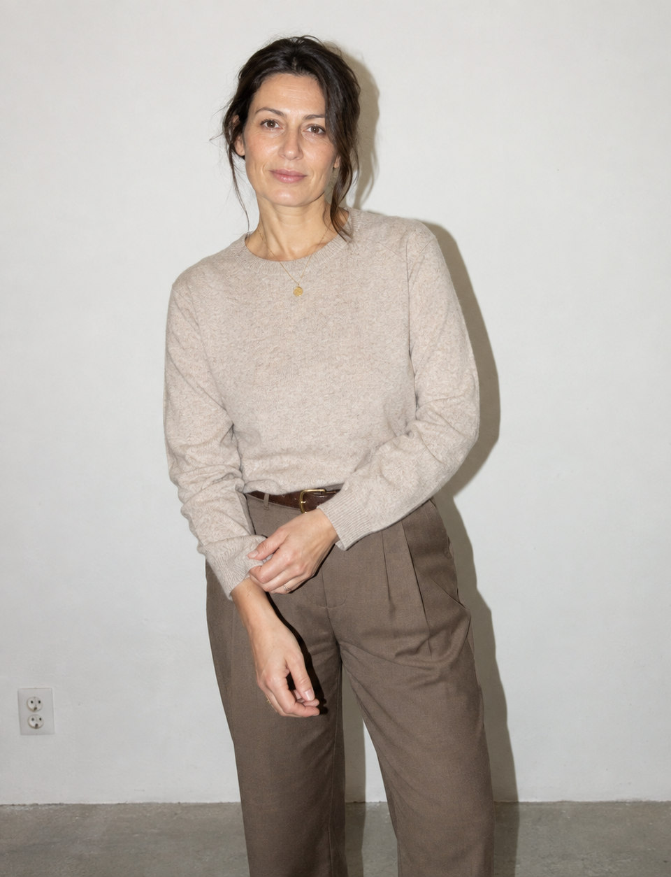 A woman stands against a plain off-white wall wearing a light beige knit crewneck sweater and brown wide-leg trousers.