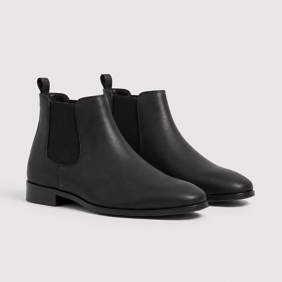 A pair of black leather Chelsea boots with black elastic side panels, shown from a three-quarters angle on a light gray studio background.