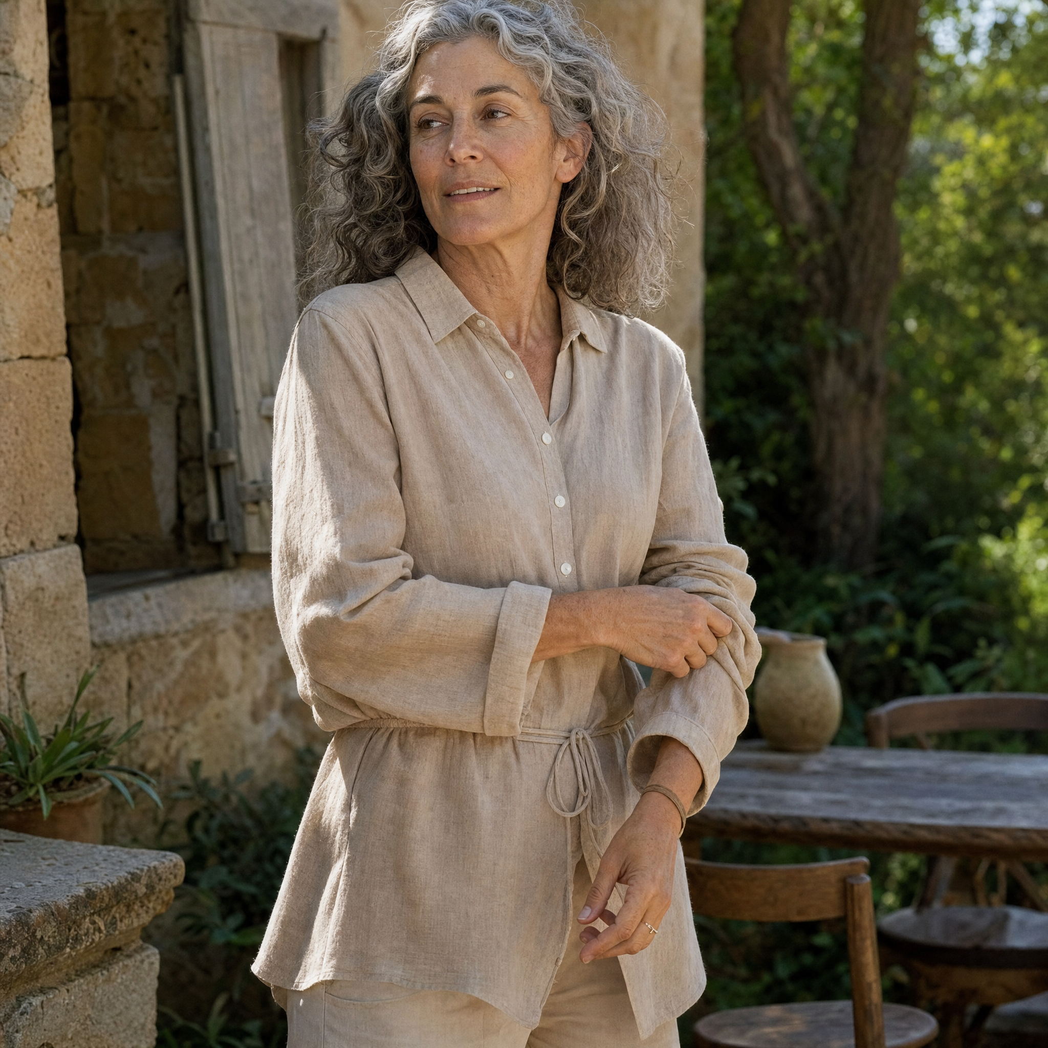 A woman with curly gray hair wearing a light beige linen long-sleeve shirt and matching pants, standing outdoors in front of a rustic stone building.